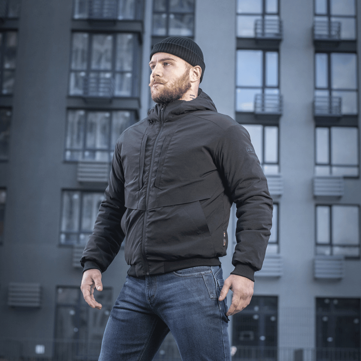 Mand iført M-Tac Rubicon Jakke Gen. II, en stilren bomberjakke til mellemsæsonen, foran en moderne bygning.