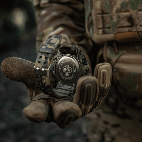 M-Tac Tactical Ur 2037 i kamuflasjedesign, vannbestandig opptil 5ATM, holdt av en taktisk hanskekledd hånd i skogsmiljø.