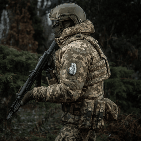 Soldat i kamuflasjeuniform med M-Tac Patch M67 Granat 3D PVC på ermet, bevæpnet i skogen.