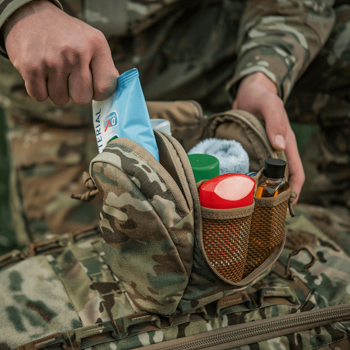 Person åbner M-Tac Toilettaske Elite Gen.II med diverse hygiejneartikler i kamuflage design, ideel til rejseopbevaring.