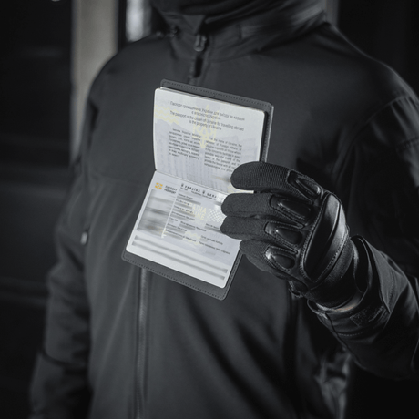 Person holding pasbeskyttelse M-Tac Pascover til ukrainske pas, visende "Passport" tryk, polymerkomposit, sort baggrund.