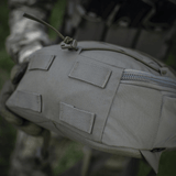 Nærbillede af taktisk rygsæk i robust materiale og beige farve, holdt af en person i camouflageuniform. Fokus på lynlås og stropper.