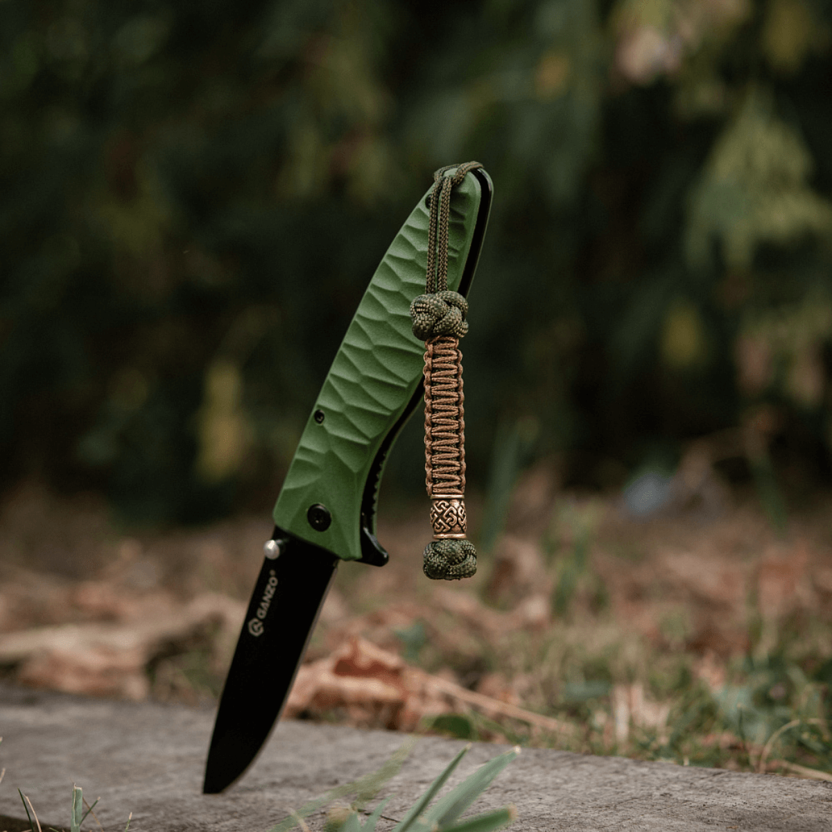 M-Tac Knife Lanyard Celtic: Flettet paracord lanyard på grøn kniv i naturlig udendørs omgivelser, stilfuldt og praktisk tilbehør.