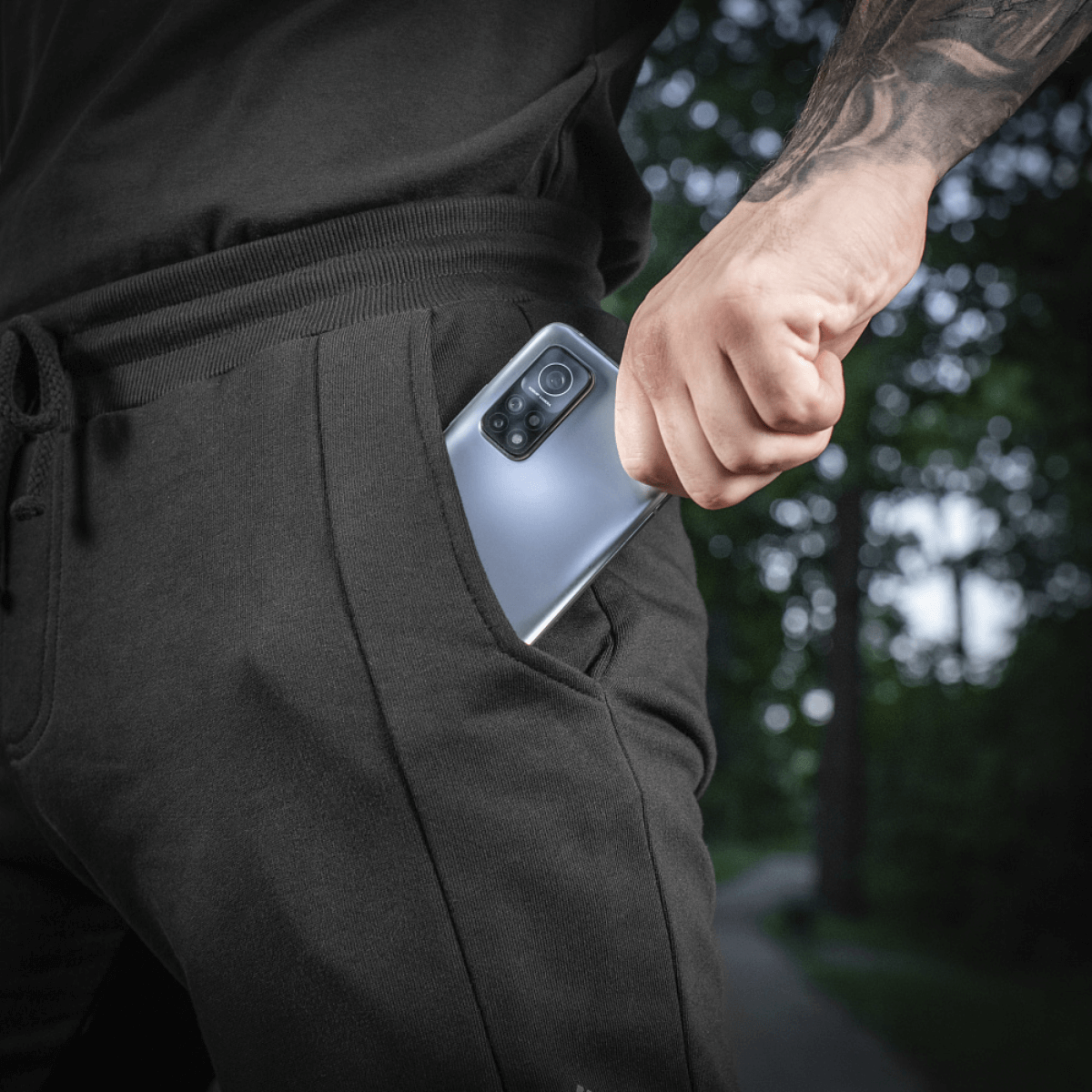 Mand iført sort joggingtøj tager en smartphone op af lommen, udendørs scene med grønne træer i baggrunden.