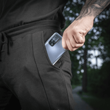 Mand iført sort joggingtøj tager en smartphone op af lommen, udendørs scene med grønne træer i baggrunden.