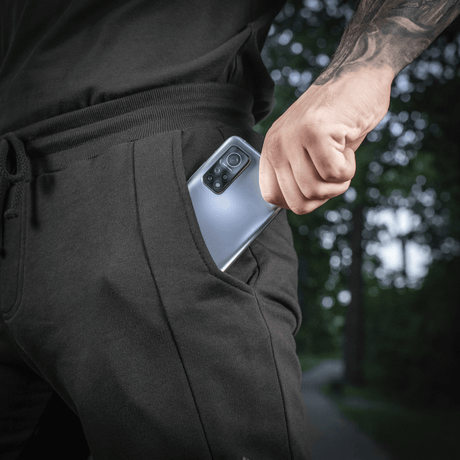 Mand iført sort joggingtøj tager en smartphone op af lommen, udendørs scene med grønne træer i baggrunden.