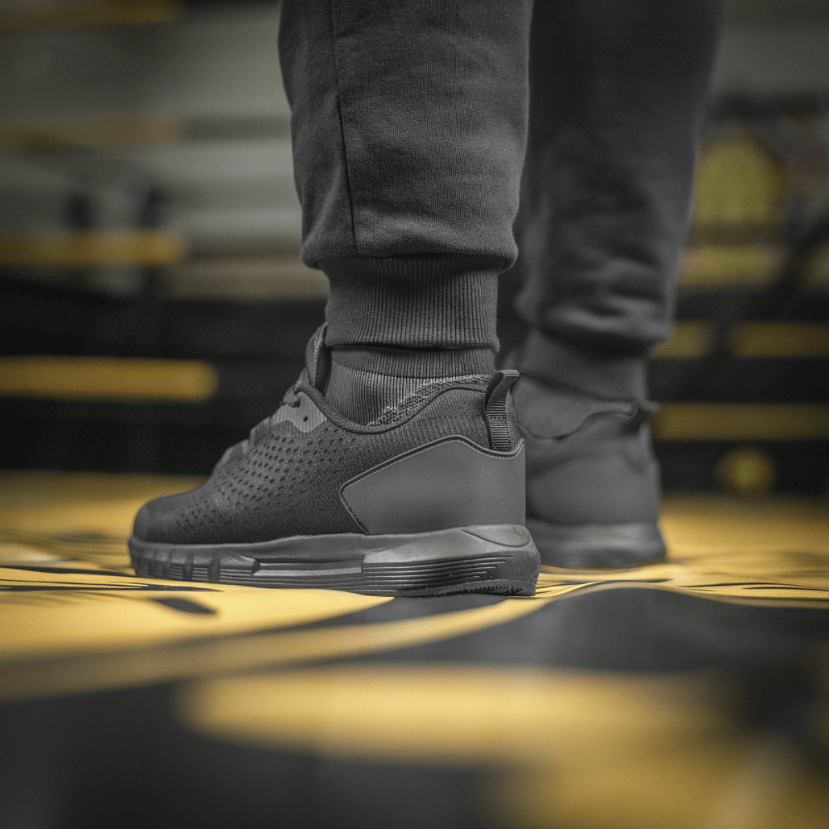Close-up of athletic shoes paired with joggers on a black and yellow patterned floor in a sports setting.