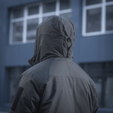 M-Tac Alpha Gen.III Pro Vinterjakke set bagfra i bymiljø, viser justerbar hætte og forstærkede områder på skuldre