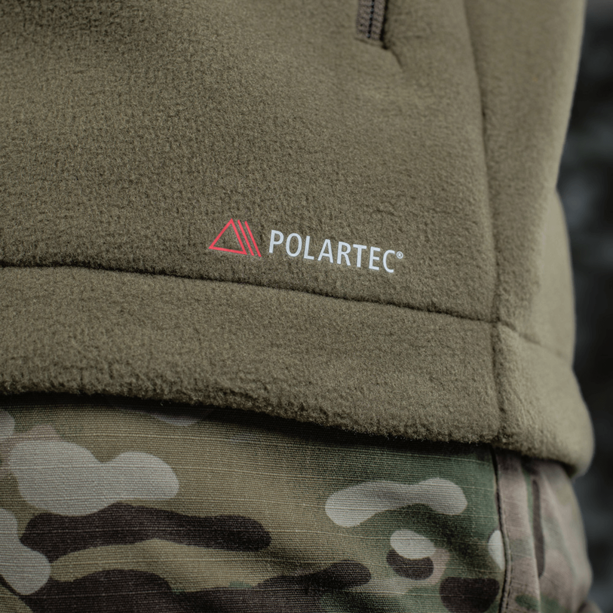 Detalje af M-Tac Combat Fleece Polartec Jakke med Polartec-logo på camouflagebaggrund, fremhævende jakken i høj kvalitet.