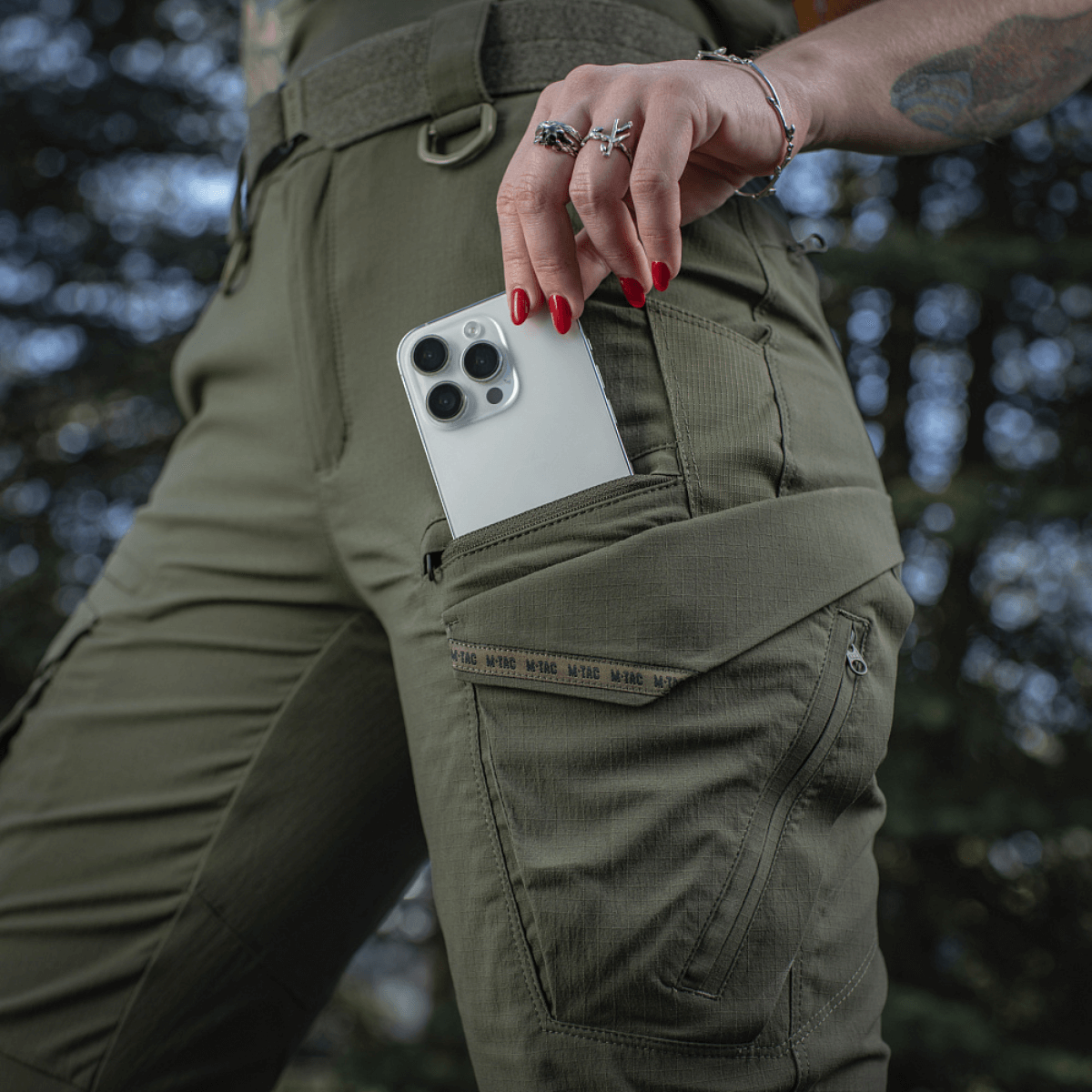 Kvinde iført M-Tac Bukser Aggressor Summer Flex Dame, viser lomme med telefon, fremhæver Rip-Stop nylon og elastisk taljebånd.