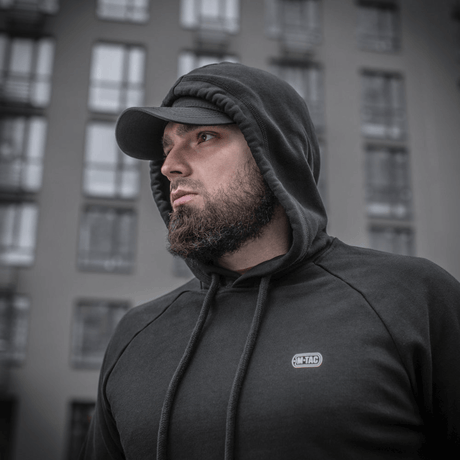 Mand iført M-Tac Cotton Raglan Hard Hoodie, 100% bomuld, med fleecefor, stilfuld til hverdagsbrug, set foran bybygning.