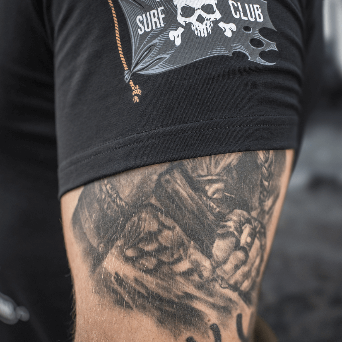 Sort M-Tac T-Shirt Surf Club med en surfkranium printdetalje og tatoveret underarm, designet til at symbolisere styrke og stil.