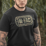 Mand iført M-Tac T-Shirt Logo i camouflage med slogan på ærmet og "Military&Tactical Since 2014" på ryggen, omringet af grønne planter.