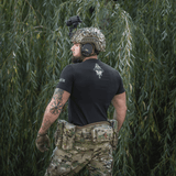 Mand iført M-Tac T-Shirt Odin Mystery foran træer, bagfra, med taktikudstyr. Fokus på figur og komfort i mytologi-inspireret design.