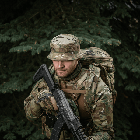 Camouflagenet mand med M-Tac Panama Hat Gen.II NYCO, taktisk uniform og rygsæk i skovmiljø.