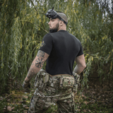 Mand i taktisk udstyr iført M-Tac T-Shirt Delivery Service med FPV-briller står i naturen, klar til aktion.
