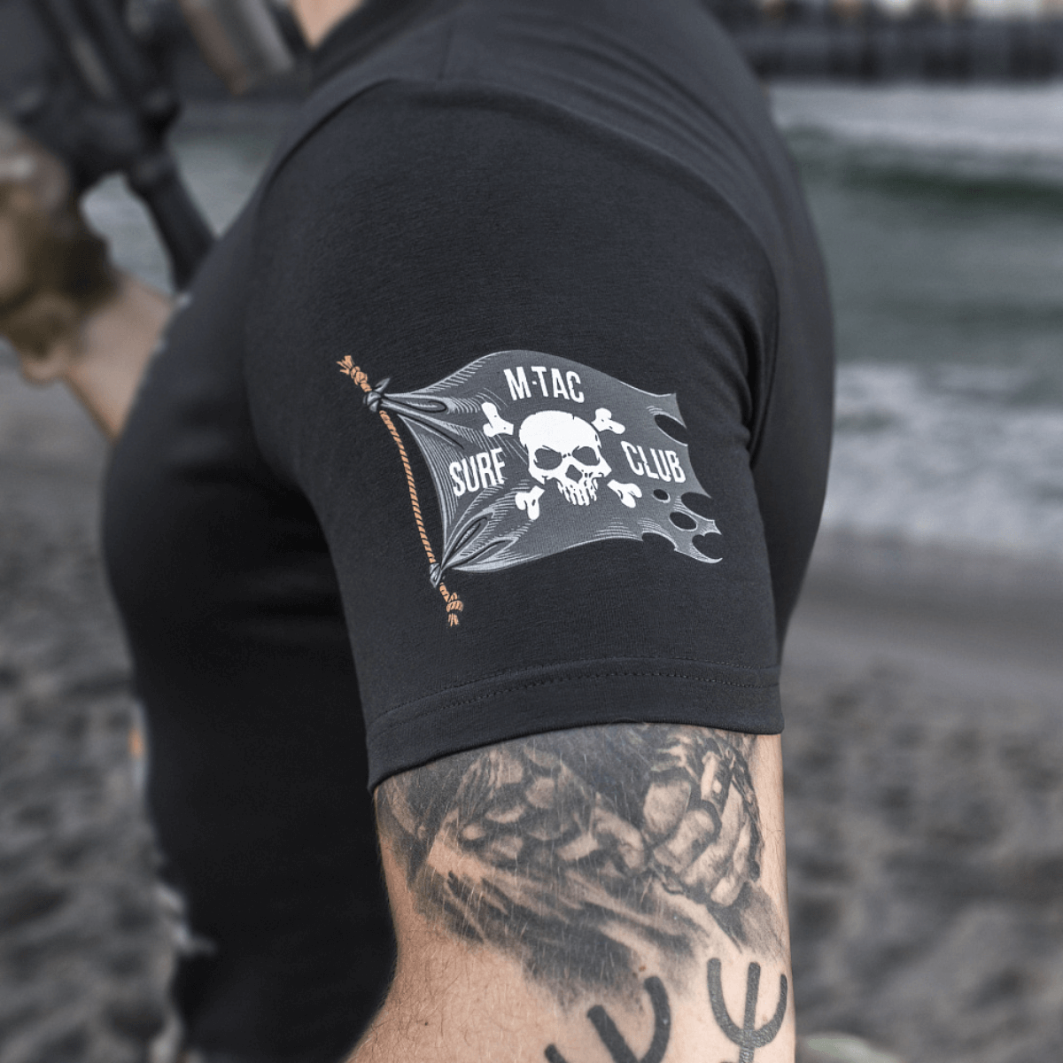 M-Tac T-Shirt Surf Club med ikonisk tryk inspireret af specialoperationer ved kysten, set på strand med soldatisk design.