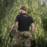 Mand bærer M-Tac T-Shirt Kamikaze Spirit med FPV-goggles og tatovering, stående i naturen.